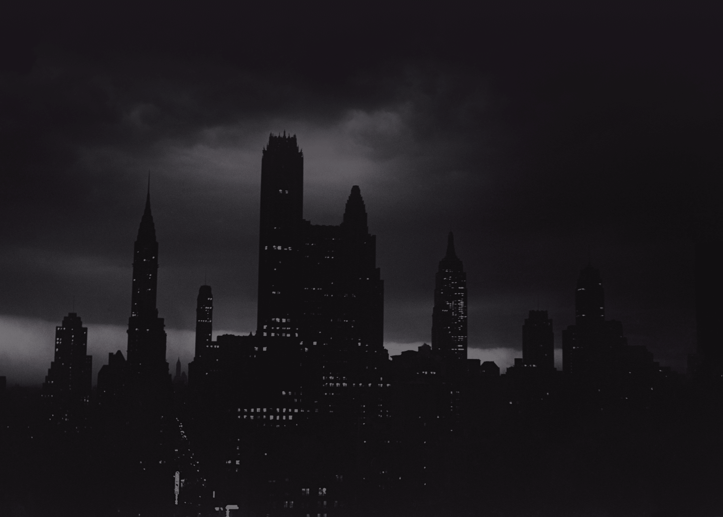 A city skyline at night with tall buildings silhouetted against a dark, cloudy sky, and scattered lights glowing from the windows