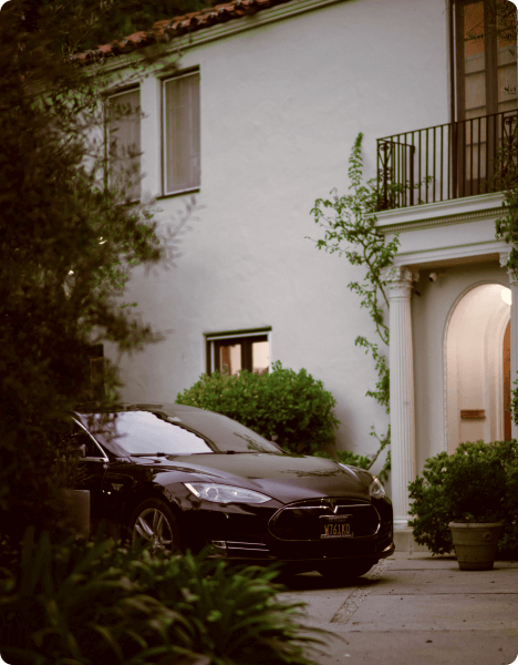 A sleek black car parked in the driveway of a two-story house surrounded by greenery during the evening.