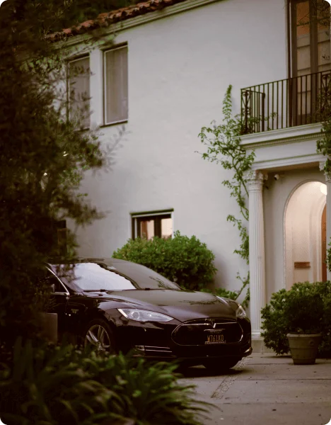 A sleek black car parked in the driveway of a two-story house surrounded by greenery during the evening.