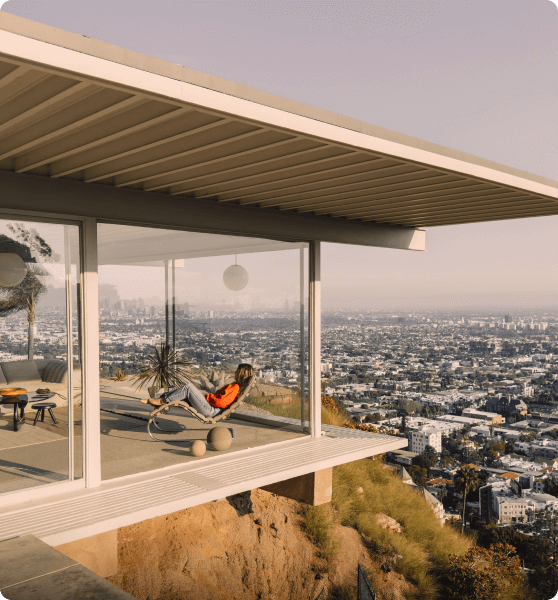 A modern house with glass walls and a person relaxing on a chair, overlooking a vast cityscape at sunset.